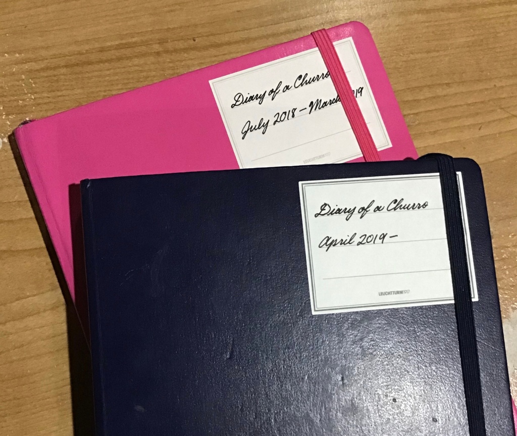 My navy journal, on top of my pink journal. The pink notebook has a sticker that says “Diary of a Churro: July 2018-March 2019” written in cursive and the navy journal has a sticker that says “Diary of a Churro: April 2019” also written in cursive.